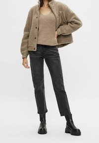 Persona che indossa un top in maglia beige, una giacca in pile tan con bottoni, jeans dritti neri e stivali con tacco spesso neri.
