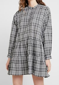 Svart och vit rutigt klänning med långa ärmar, knäppning framtill, lager-på-lager kjol och lätt material. Har en rund halsringning och rynkade detaljer.