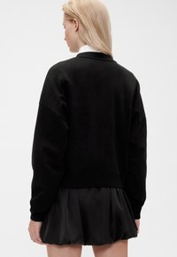 Sudadera negra con un corte relajado, cuello redondo y puños acanalados, combinada con una falda negra plisada. Textura suave y diseño corto.