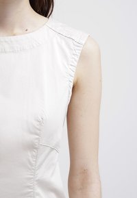 Ljusfärgad ärmlös topp med en slät yta, rund halsringning och subtila sömnadsdetaljer på axlarna; figurnära passform.