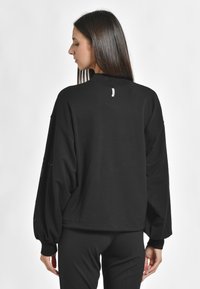 Pulover negru supradimensionat, cu mâneci lungi și puffate și guler înalt. Textură netedă, design minimalist, cu un mic eticheta pe spate.