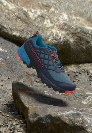 Μπλε και μαύρο παπούτσι για τρέξιμο σε μονοπάτια με πορτοκαλί λεπτομέρειες, αιωρούμενο πάνω από πετρώδη έδαφος, με ανθεκτική σόλα και λογότυπο La Sportiva.