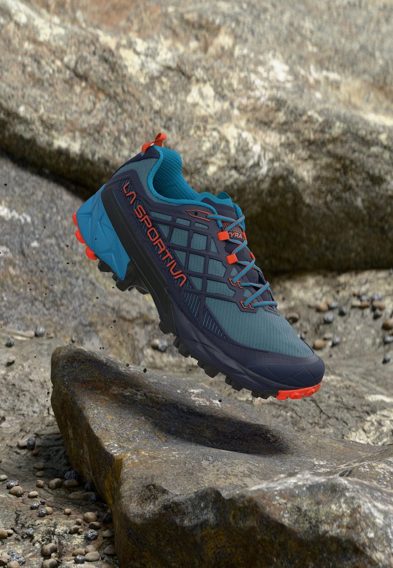Μπλε και μαύρο παπούτσι για τρέξιμο σε μονοπάτια με πορτοκαλί λεπτομέρειες, αιωρούμενο πάνω από πετρώδη έδαφος, με ανθεκτική σόλα και λογότυπο La Sportiva.