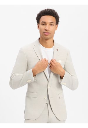Młody mężczyzna poprawiający jasnobeżową marynarkę garniturową na białej koszulce, stojący na jednolitym białym tle.