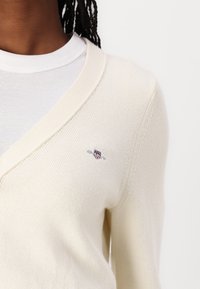 Cardigan beige a maglia con scollatura a V, dotato di un piccolo logo ricamato sul lato sinistro. Il materiale appare morbido e texture.
