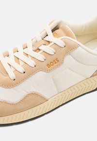 Baskets à lacets en daim beige et cuir lisse blanc, avec une semelle en caoutchouc beige texturé et un logo "BOSS" en doré.