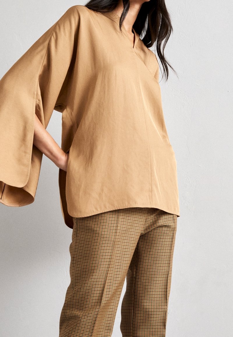 Blouse beige oversize avec un col en V et des manches larges, associée à un pantalon marron à carreaux, mettant en valeur un tissu texturé et une coupe décontractée.