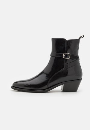 Botín de cuero negro con un acabado brillante, punta afilada y un tacón bajo en bloque. Presenta un detalle de tira decorativa con hebilla.