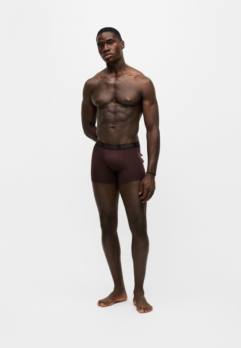 Homme musclé debout pieds nus, bras détendus, portant un boxer Puma marron foncé sur fond blanc uni.
