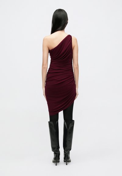 Bordó egyrészes ruha, gyűrt textúrával, testhez simuló fazonnal és aszimmetrikus szegéllyel. Fekete, térdig érő tűsarkú csizmával párosítva.