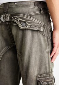 Pantalons cargo en denim olive délavé, avec une finition texturée, des clous en métal sur la ceinture et une poche latérale zippée.