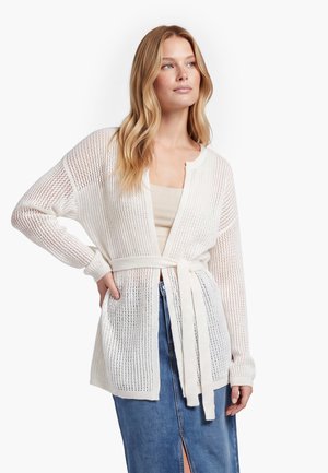Kvinden bærer en hvid åbenstrikket cardigan med bælte, beige top og blå denimnederdel med et frontslids og poserer med den ene hånd på hoften.