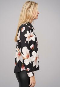 Zwarte blouse met grote witte en roze bloemenprint, lange mouwen, een kraag en een ontspannen pasvorm. Bodem lengte tot op de heup. Gladde stoftextuur.