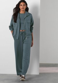 Two-piece loungewear set in muted teal; includes a zippered jacket with pockets and loose-fitting pants with an adjustable waistband. Soft fabric.