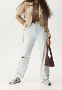 Veste de motard en daim beige avec zips, chemise rayée marron et verte, jean taille haute bleu clair déchiré, baskets grises et blanches, sac marron.