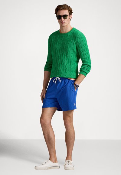 Πράσινο πουλόβερ με πλέξη καλώδιο, μπλε σορτς με κορδόνι, λευκά αθλητικά παπούτσια, γυαλιά ηλίου και ρολόι. Περιλαμβάνει πορτοκαλί λεπτομέρειες στα σορτς.