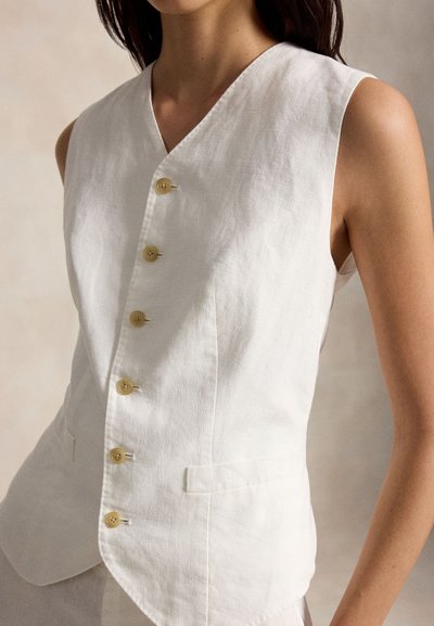 Αμάνικο λευκό γιλέκο από λινό με έξι ανοιχτό καφέ κουμπιά και εμπρόσθιες τσέπες, φορεμένο από ένα άτομο με σκούρα μαλλιά πάνω σε ουδέτερο φόντο.