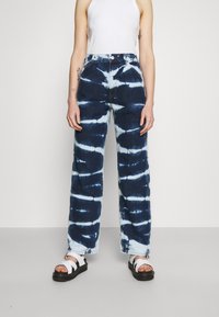 Blå batikfärgade jeans i denim med hög midja, vida ben och kontrasterande vita mönster. Bärs med vita sandaler, visade mot en neutral bakgrund.