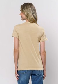 Béžové tričko s krátkým rukávem z lehké látky, s klasickým kulatým výstřihem a volnějším střihem, doplněné světle modrými džínami.