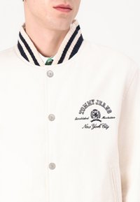 Hvit jakke med krage i marineblå og hvite striper, sølv snapknapper og brodert "Tommy Jeans"-logo på venstre bryst.
