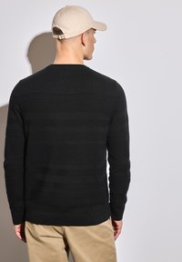 Pull en laine noir à texture avec des rayures horizontales, doté d'un col rond et de manches ajustées, associé à un pantalon kaki et une casquette beige.