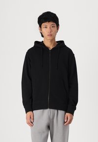 Sudadera con cremallera negra hecha de una tela suave, con capucha forrada, bolsillos frontales y puños acanalados. Usada con pantalones de chándal gris claro.