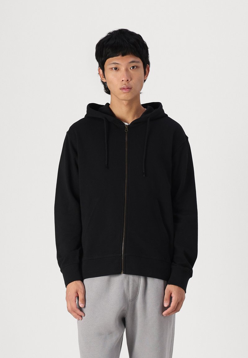 Sudadera con cremallera negra hecha de una tela suave, con capucha forrada, bolsillos frontales y puños acanalados. Usada con pantalones de chándal gris claro.