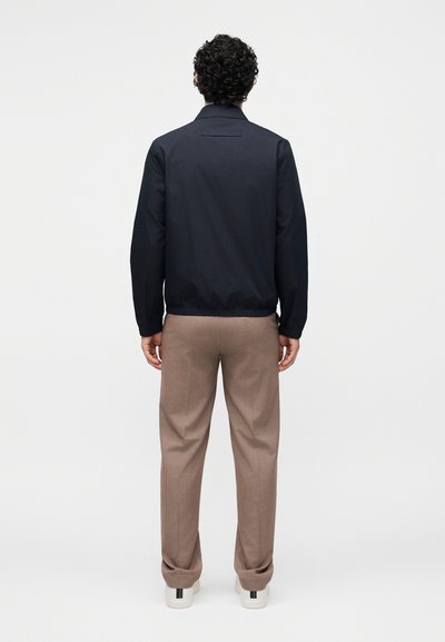 Άνδρας με κατσαρά μαλλιά, φοράει μαύρο μπουφάν, μπεζ παντελόνια και λευκά αθλητικά παπούτσια, στέκεται με την πλάτη σε απλό ανοιχτό φόντο.