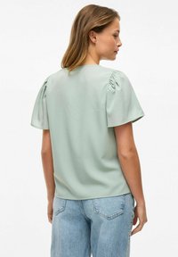 Svijetlozelena bluzica kratkih rukava s napučenim rukavima, glatke teksture i okruglim izrezom, uparena s trapericama visokog struka u plavoj boji.