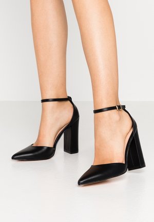Black leather high-heeled shoes with a pointed toe, chunky heel, and ankle strap featuring a gold buckle detail. Smooth texture, open sides.