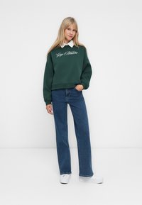 Woman standing wearing a dark green sweatshirt with "Gap Athletic" text, blue jeans, white sneakers, and a white collared shirt underneath.