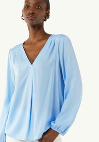 Hellblaue Bluse mit V-Ausschnitt, langen Ärmeln und gerafften Manschetten. Hergestellt aus glattem Stoff mit lockerem Schnitt und subtilen Faltendetails.