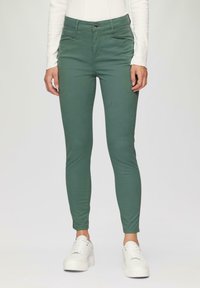 Grüne, schmal geschnittene Hose aus glattem Stoff, mit einem standardmäßigen Reißverschluss und Fronttaschen, kombiniert mit weißen Sneakers.