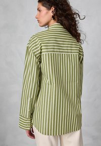 Žena s kudrnatými vlasy, která nosí zlaté geometrické náušnice a olivově zelenou dlouh sleeve košili s vertikálními bílými pruhy, zobrazená zezadu.