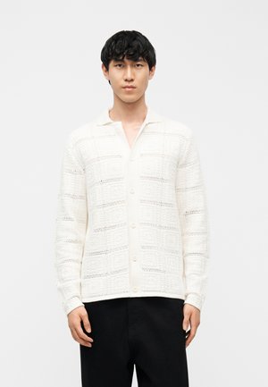 Fehér, gombos kardigánt viselő férfi, amelyen texturált négyzetmintázat található, fekete nadrágban, egyszínű fehér háttér előtt áll.