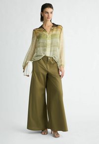 Blouse transparente vert clair à rayures horizontales, associée à un pantalon large olive. Les accessoires comprennent une pochette blanche et des sandales à motifs.