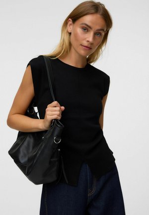 Femme aux cheveux châtain clair portant un top sans manches noir et un jean bleu foncé, tenant un sac à main en cuir noir sur son épaule.