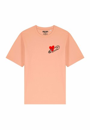 T-shirt color pesca con maniche corte, caratterizzato da una spilla di sicurezza con contorno nero e un grafico di un cuore rosso nell'area sinistra del petto.