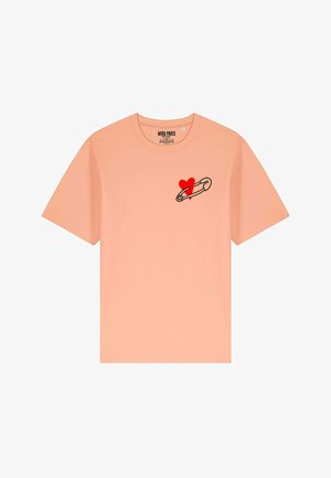 T-shirt color pesca con maniche corte, caratterizzato da una spilla di sicurezza con contorno nero e un grafico di un cuore rosso nell'area sinistra del petto.