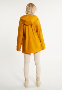 Abrigo impermeable amarillo con capucha, que presenta un corte relajado, puños abotonados y una textura suave. Combinado con pantalones ajustados de color claro.