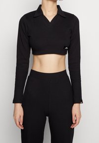Schwarzes geripptes Langarm-Crop-Top mit Kragen und V-Ausschnitt; verfügt über ein kleines Logo-Patch auf der linken Seite. Texturiertes Material.