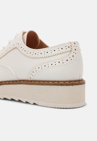 Chaussure en cuir blanc avec une surface texturée, des surpiqûres décoratives et des accents perforés. Dispose d'une semelle claire, rainurée, avec un léger talon.