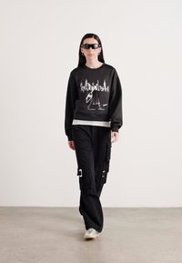 Sweatshirt preto com uma impressão gráfica de um horizonte urbano; combinado com calças cargo pretas com detalhes de fivela e tênis.
