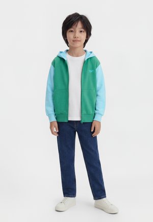 Barn iført en grøn og lyseblå hættetrøje med lynlås, hvid t-shirt, blå jeans og hvide sneakers står mod en ensfarvet baggrund.
