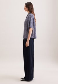 Femme aux longs cheveux bruns vue de profil, portant un haut ample à manches courtes à motifs et un pantalon noir à jambes larges avec des chaussures noires à bout pointu.