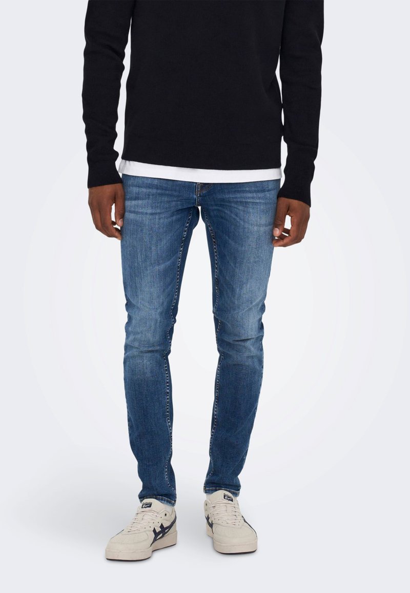 Jeans skinny albastre cu un aspect estompat, asortați cu o bluză neagră cu mânecă lungă și un top alb în straturi. Purtați cu adidași bej.