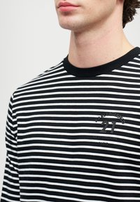 Közeli kép egy fekete-fehér csíkos pólót viselő személyről, akinek a mellkasán "KARL LAGERFELD 21 RUE ST-GUILLAUME PÁRIS" felirat látható.