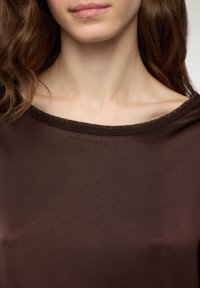 Top marrone con una ampia scollatura a coste arricchita da accenti metallici; il tessuto ha una texture liscia e leggermente lucida.