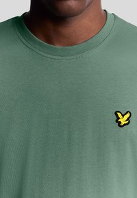 Maglietta in cotone verde con scollo rotondo e maniche corte, caratterizzata da un logo di un uccello nero e giallo sulla parte sinistra del petto.