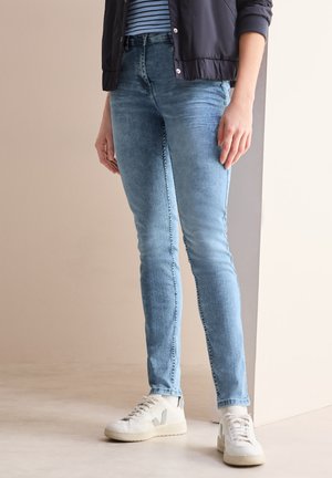 Jeans skinny bleu clair avec une texture délavée, taille haute et coutures contrastées, associés à des sneakers blanches et une chemise rayée.